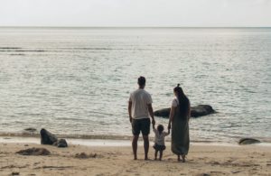 Family of three standing on a beach by the ocean.