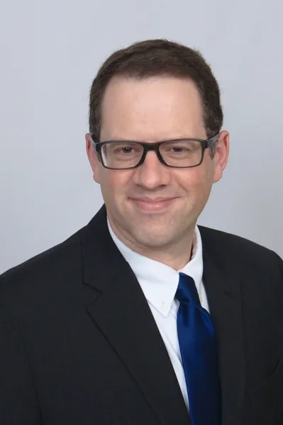 Portrait Image of Brian Burke immigration lawyer. Brian is wearing a black business suite, white shirt, and a dark blue tie. Brian is in front of a dark gray background.
