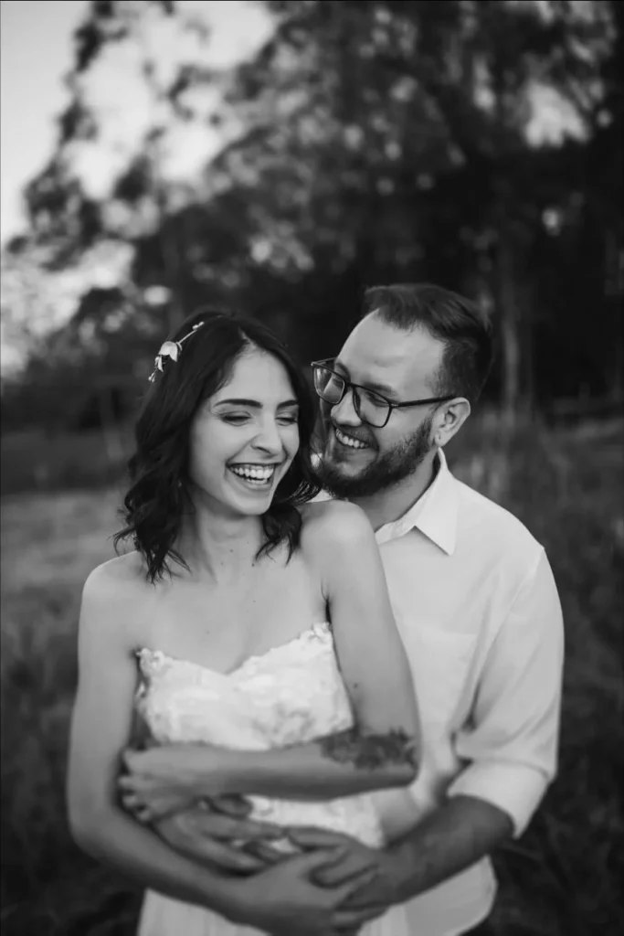 Image of wife and husband in a park. They are hugging and the husband is kissing the wife on the cheek while holding her from behind. She's wearing a white wedding dress and the man is wearing a white collar shirt. The picture is in white and black and they are at a park with trees. The image is a representation of the K-3 Spouse Visa Immigration lawyers of Shihab Burke LLC
