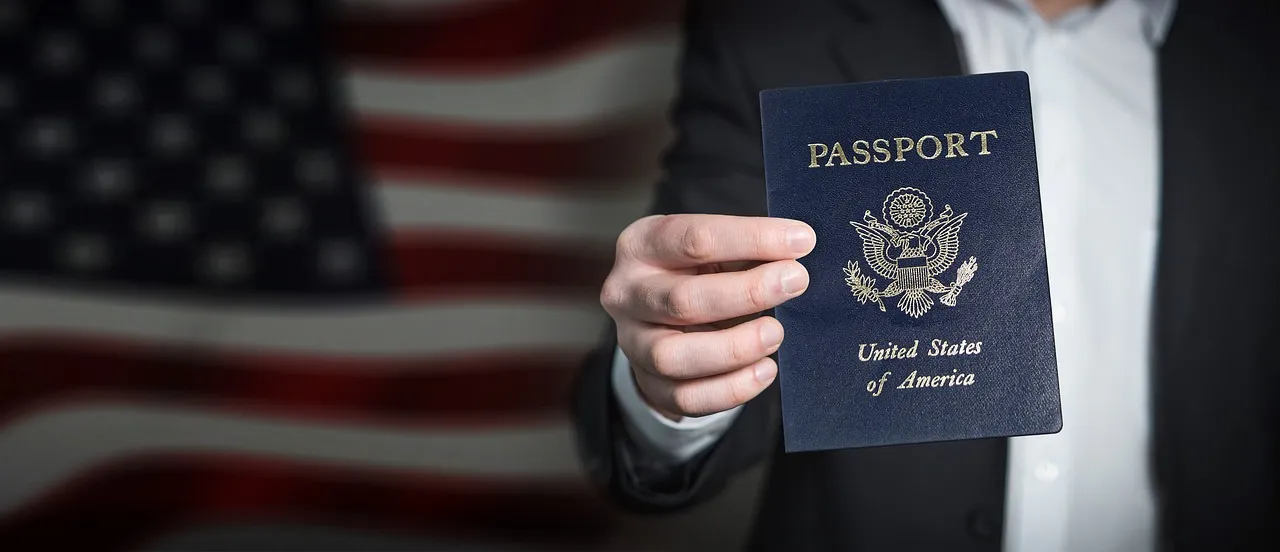 Image of an Attorney is holding an American passport up in front of him, wearing a business suit, and he is standing in front of a large American flag which is mounted on wall. The image represents the American work visas attorney at Shihab Burke LLC Immigration Lawyers.