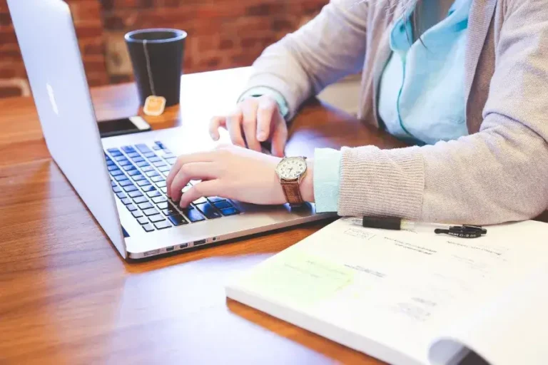 Image of person working on a desk with their computer, some paper documents, and a cup of tea. The person is wearing a sweater, and light brown watch, and a sky blue long sleeved work t-shirt. The photo represents the EB-1 Visa Extraordinary ability immigrants who apply for the EB-1 Visa.