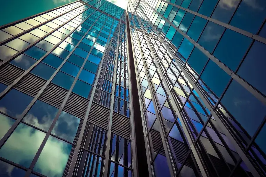 Image of a skyscraper. The angle is shot from the bottom floor, pointed up toward the sky and the skyscraper's glass windows. The glass windows reflect the cloudy sky outside. The image represents the Outstanding Professors and Researchers who apply for the EB-1 Visa.