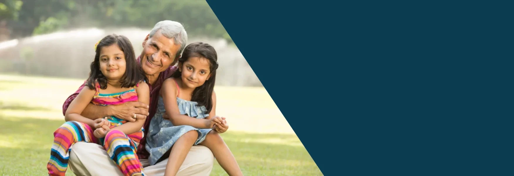 This is a banner image with half its right section colored in dark teal color and the other half there is an image of a grandfather hugging his granddaughters in a park. In the background you see green leafy trees. This is a representation of family unison through immigration law.