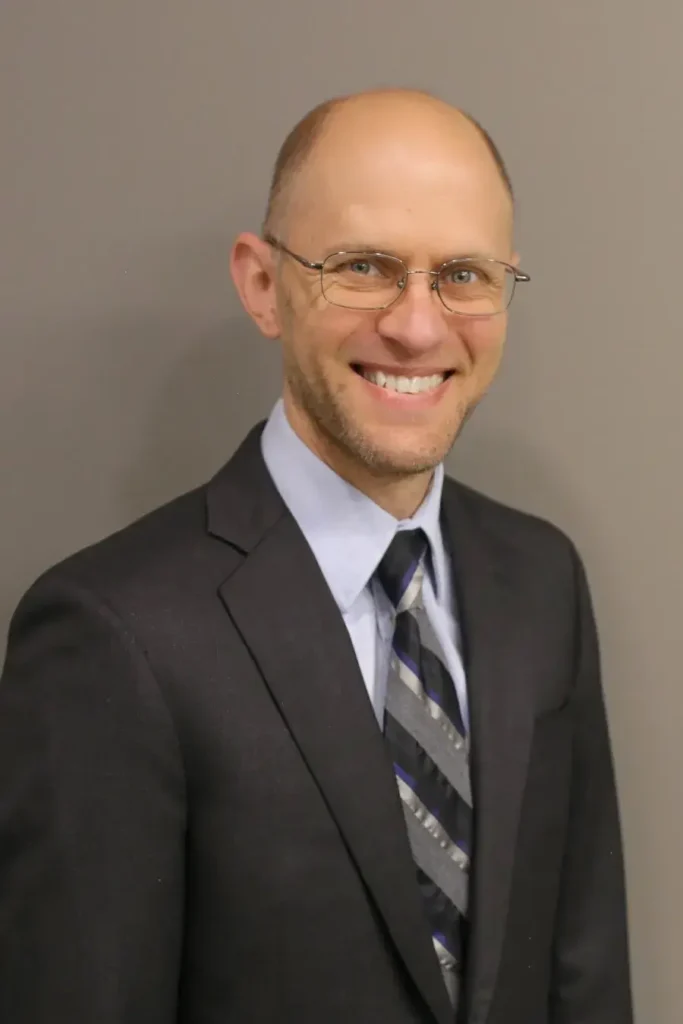 Portrait Image of Joe Morehart immigration lawyer from Shihab Burke LLC Immigration Lawyers. Joe is wearing a dark grey business suite, light blue business shirt, and a black, white, grey striped tie. Joe is in front of a dark beige backdrop.