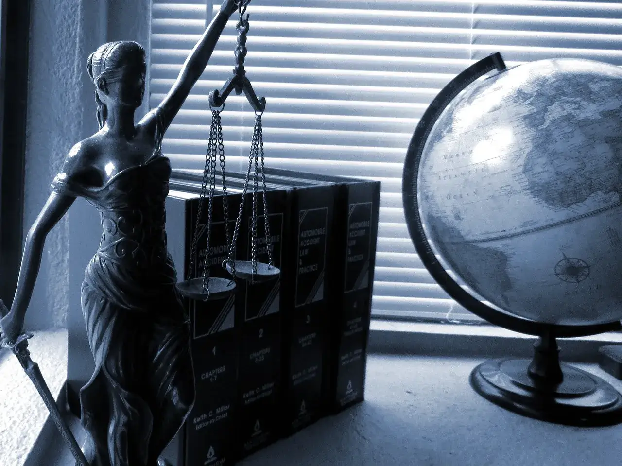 Image of a Lady Justice miniature statue placed on top of a desk. There is also a mini globe next to a set of books that are also on top of the same desk. In the background there is a window with closed blinders. There image is in black and white. The image is used to represent the PERM Labor Certification Lawyers of Shihab Burke LLC