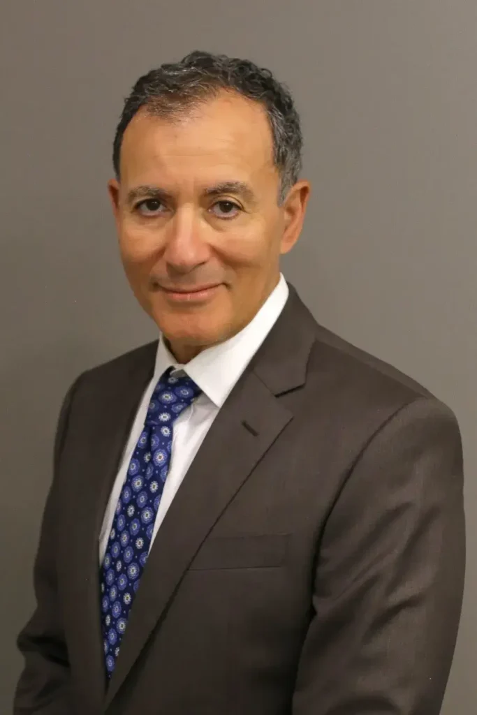 Portrait Image of Sam Shihab immigration lawyer from Shihab Burke LLC Sam is wearing a dark grey business suite, white business shirt, and a blue tie with white diamond printed design. Sam is in front of a dark beige backdrop.