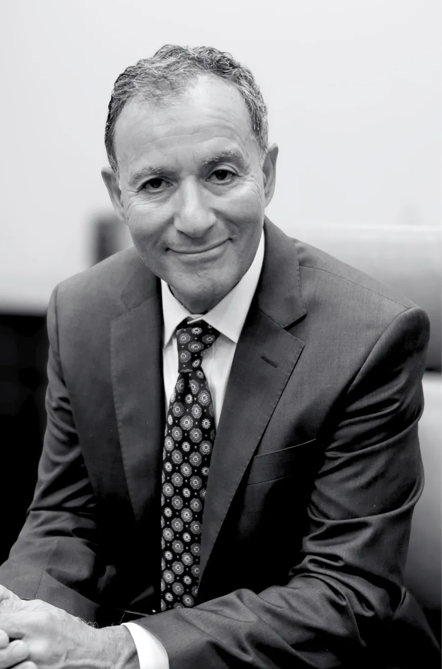 Portrait Image of Sam Shihab from the Shihab Burke LLC Immigration Lawyers law firm. Sam is sitting with his hands in front folded atop a desk. He is angled slightly and his head head turned toward the camera. He is smiling. The picture is in black and white.