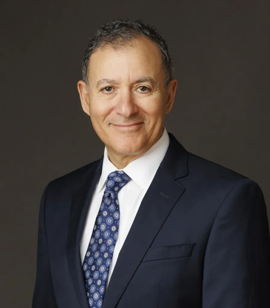 Portrait Image of Sam Shihab from the Shihab Burke LLC Immigration Lawyers law firm. Sam is sitting with his hands in front folded atop his lap. He is angled slightly and his head head turned toward the camera. He is smiling and wearing a blue patterned tie.