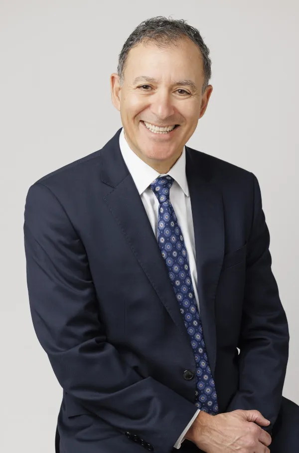 Portrait Image of Sam Shihab from the Shihab Burke LLC Immigration Lawyers law firm. Sam is sitting with his hands in front folded atop his lap. He is angled slightly and his head head turned toward the camera. He is smiling.